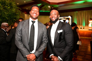 Two smiling men in formal suits posing for a photo at a black-tie event, with green uplighting in the background.
